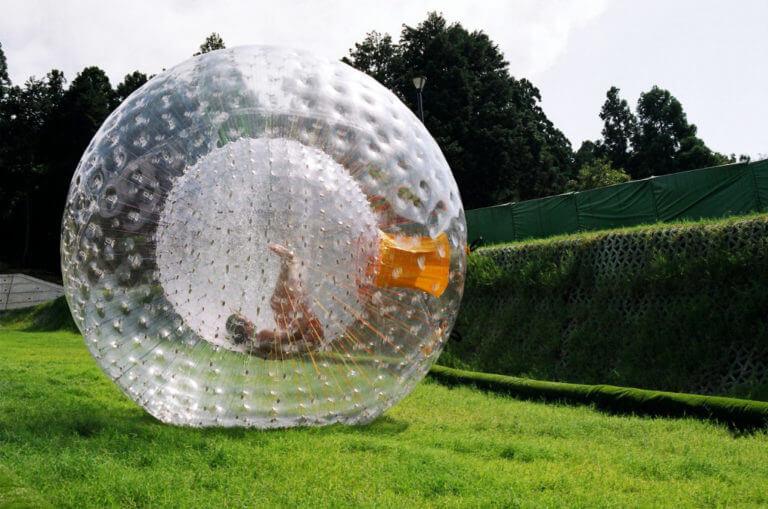 KALNU ZORBING RIGA. DIVI CILVĒKI TRĪS METRUS GARĀ GAISA BALONĀ, KAS SLĪD NO KALNA.
